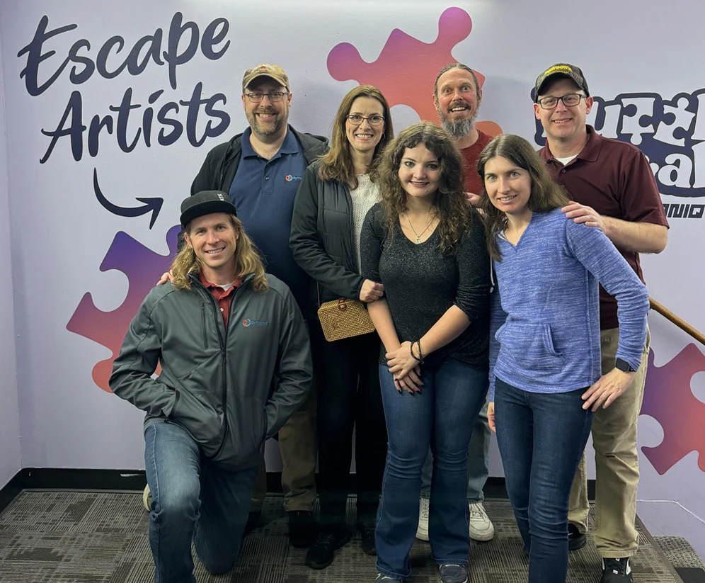 Seven members of the dymaptic Software Development team posing in front of an Escape Artists puzzle-themed mural after completing an escape room in Seattle, WA, with several team members wearing dymaptic-branded jackets.
