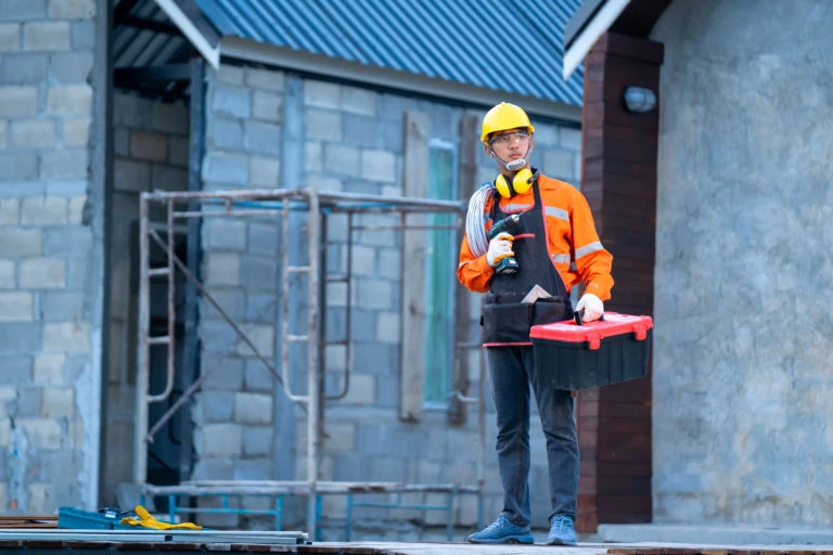 A construction worker in an orange safety jacket and yellow hard hat stands on a job site holding a toolbox and power drill, used to illustrate the concept of having the right tools for the job.