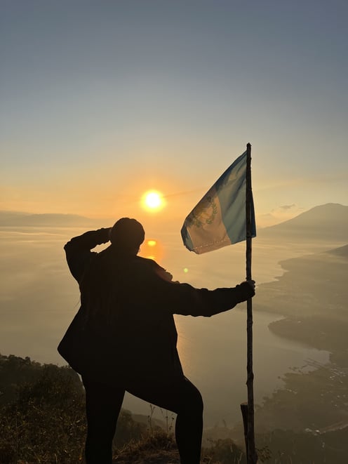 A photo of a woman, holding a Guatemalan flag, overlooking a shoreline, with the sun low on the horizon
