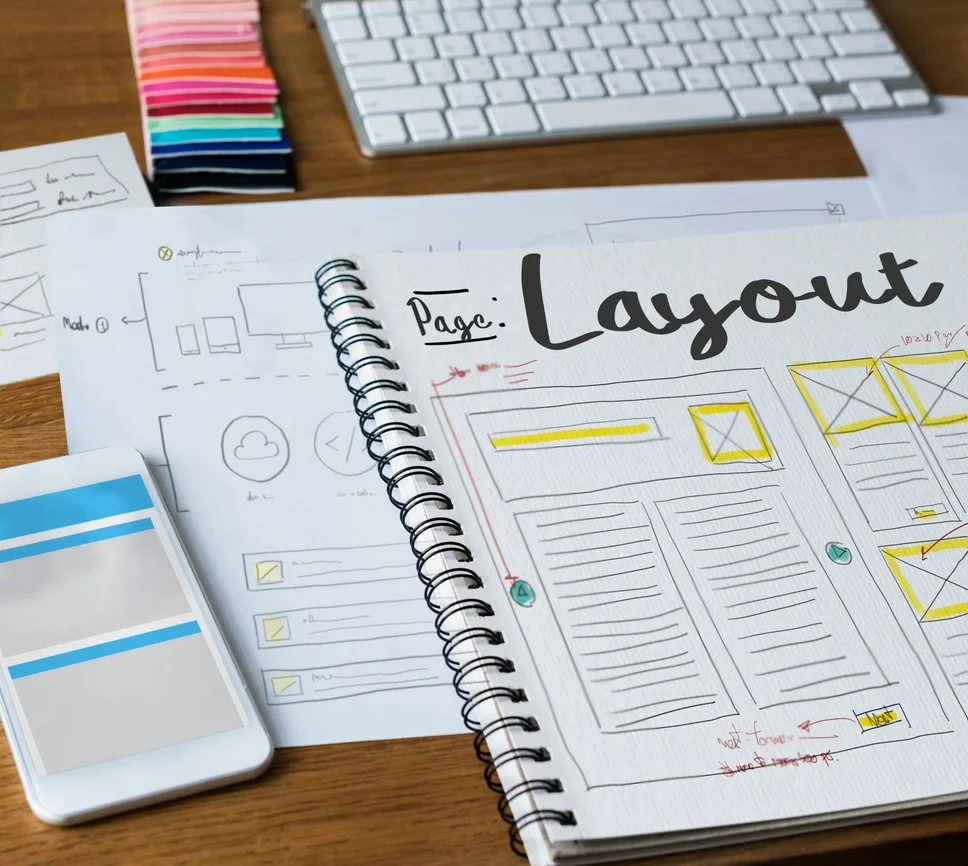 A designer's desk with a spiral notebook open to hand-drawn webpage wireframes labeled Page: Layout, surrounded by printed interface sketches, a color swatch fan, a keyboard, and a smartphone mockup, illustrating the planning and wireframing phase of web design.