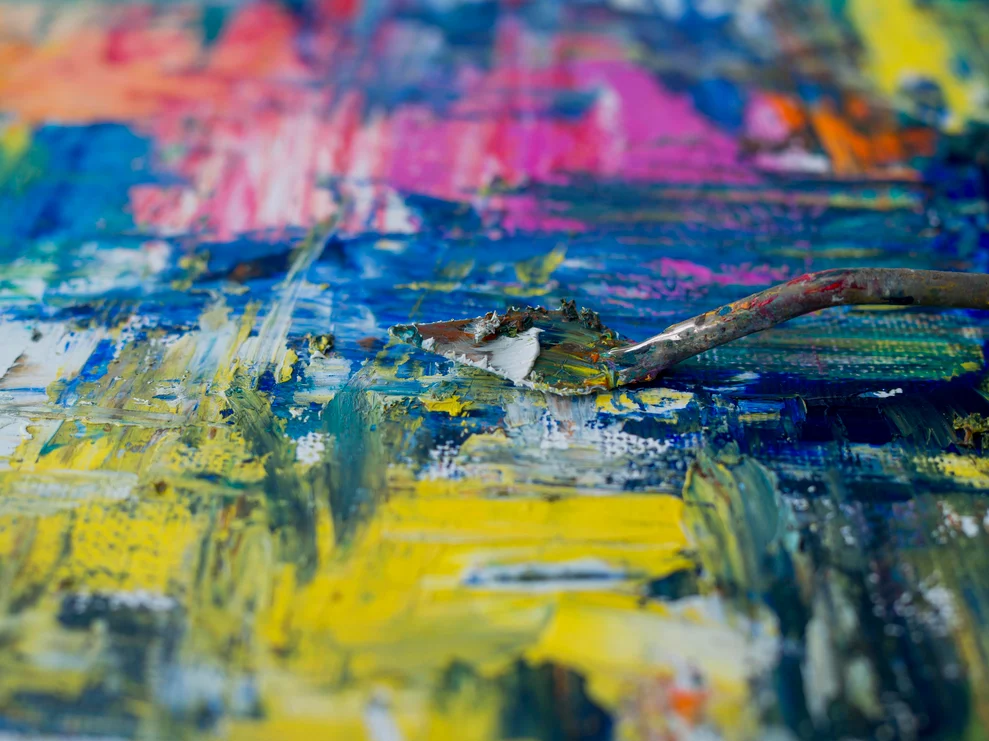 Close-up of a palette knife resting on a heavily textured canvas covered in bold brushstrokes of blue, yellow, pink, and green oil paint, illustrating the tactile texture of a painting in progress.